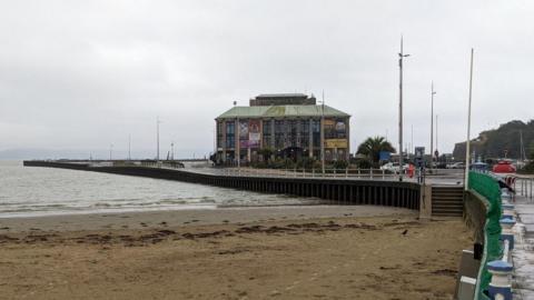 Weymouth Pavilion and peninsula from the Esplanade on a grey day. The peninsula is almost entirely a car park with the large green-roofed Pavilion building in the centre.