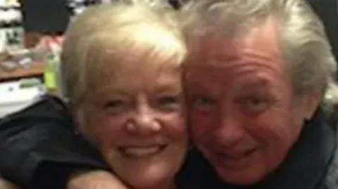 Photograph of Sheila Jackson, who has blonde hair, and Eric Greener, who has grey hair. Mr Greener has his arm around Ms Jackson's shoulder and they are both smiling for the camera in the close-cropped shot. 