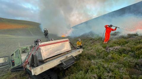 A tracked vehicle and two people in protective overalls and hard hats tackle flames burning in a large area of moorland in the Cairngorms.