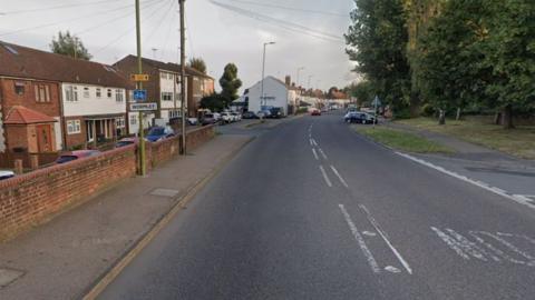 Wormley High Road at one end. There is a pavement on the left bordered by a low brick wall and terraced houses behind it which continue into the distance. The right side of the road has a grass verge, pavement behind it and some green trees. 