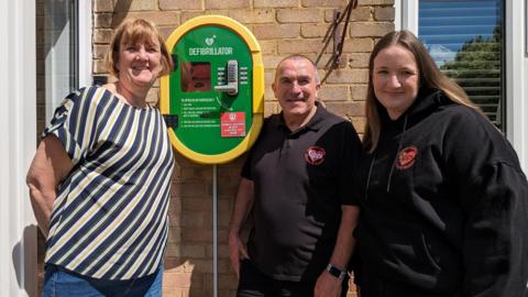 A woman in a striped top, a man in a branded polo shirt and a woman wearing a hooded top with the same logo stand next to a defibrillator unit and smile.