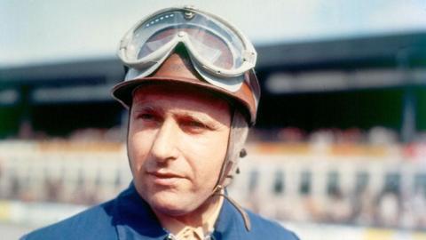 Juan Manuel Fangio wearing a blue racing suit and brown helmet with googles.