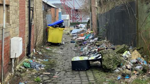 Rubbish filling an alleyway. Full bins, a suitcase, pillows, and litter are strewn across the length of the path.