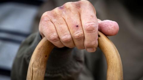 A close-up of the hand of an elderly man holding a wooden walking stick.