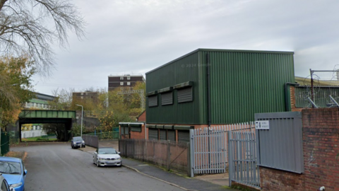 A street with green and redbrick warehouse buildings on the right side, behind a metal fence. A railway bridge can be seen towards the end of the street.