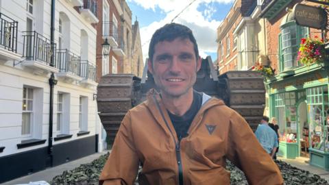A man with short dark hair wears a black t-shirt and bronze coloured jacket. He is sitting on the back of lorry which is carrying a vintage tank. The vehicle is travelling through a town centre street lined with historic buildings.