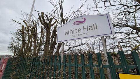 The White Hall Academy sign outside the school amongst some bramble and behind a green fence