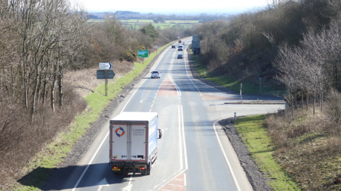 A lorry turning right on a section of the trunk road. It is viewed from above and there are other vehicles in the distance and trees up either side of the road.