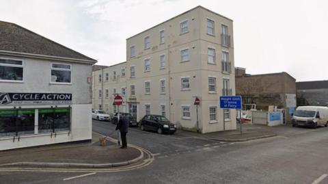 A Google Street View of the junction of Quarry Street and Harvey Street in Torpoint. A man is stood on the pavement looking down Quarry Street on the left of the image while walking a dog. A bike shop called Cycle Action is on the corner of the junction. A cream-coloured building is on the other side of the road. A blue sign saying "Weight limit 17 tonne at Ferry" is on the road. Several cars and a white van are parked on the roads.