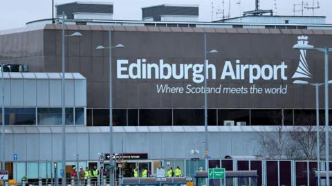 The exterior of Edinburgh Airport, the airports logo is visible in the front. There are some people wearing hi-vis outside.