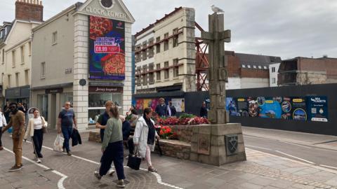 People walking past Charing Cross in St Helier, Jersey. A cross statue is on a plinth on the street. A building site with hoarding around its edge is on the opposite side of the road from the cross. A large video screen on a building called the Clock Tower has an advert for Domino's pizza on it.