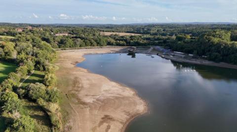 An aerial shot shows a reservoir surrounded by green fields and trees, but with large areas of brown around the edges of the reservoir where the water has receded.