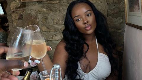 A woman wearing a white top and has her hair down is sat enjoying a glass of Prosecco. She is clinking her glass with a mysterious male looking hand sat to the other side of her.