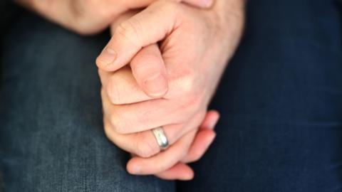 Two hands clasped together. On is wearing a silver wedding band.