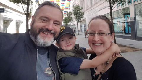 A man takes a selfie with a woman and a young boy. They are all smiling as they stand on a high street with multi-coloured umbrellas displayed above them. Several trees and a Primark store can be seen behind them. 
