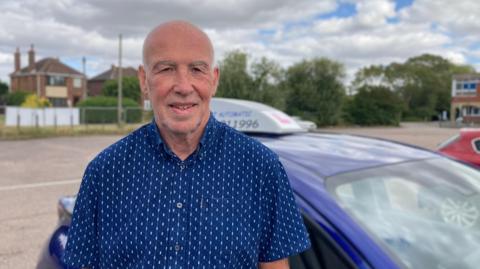 Stephen Pearson standing by the side of his blue driving school car wearing a blue shirt with white speckles. He has a bald head with grey eyebrows