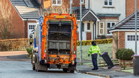 A worker in a hi-vis jacket drags a wheelie bin towards a bin lorry