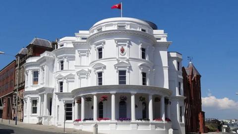 The all-white Tynwald building, which is known as the wedding cake building, which has columns at the ground floor semi-circle shape, adorned with hanging flower baskets.