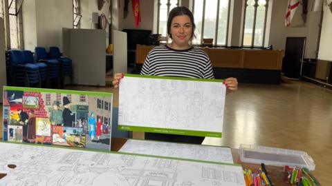 A person stands in a spacious room with arched windows and flags on the walls, holding up a partially coloured poster. Several other posters and colouring materials are spread out on the table in front of them.
