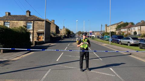 A policeman stands behind some police tape, stretched across a major road