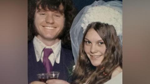 The image appears to be an older photograph of two people at a wedding. One person is wearing a bridal veil and headpiece, while the other is dressed in a suit with a wide-collared shirt and tie. Both are holding small glasses, possibly for a toast. The style of clothing and photo quality suggest it may have been taken several decades ago.