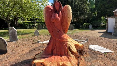 A carved wooden angel made from the stump of a tree. The angel has an orange coloured tan caused by preserving oil. Around the angel are various gravestones with more trees in the background.