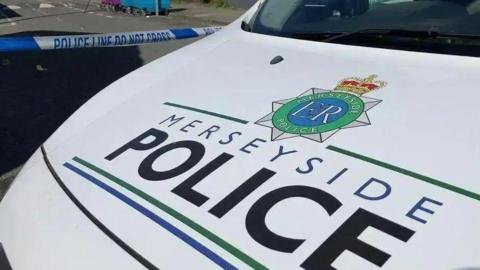 Merseyside Police vehicle parked in front of a blue and white police tape cordon. 