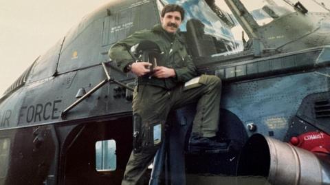 A man is standing on the side of a helicopter. He is dressed in uniform and is holding a helmet. He has dark hair and a moustache and is smiling at the camera.