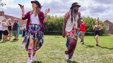 A man and a woman folk dancing on a grassed area. They are wearing colourful clothing. 