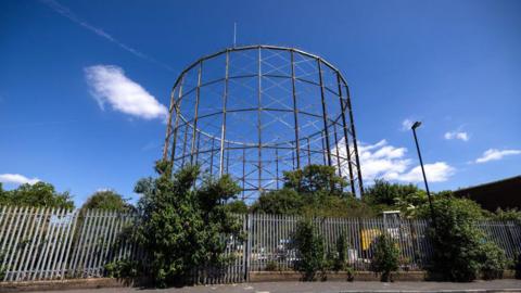 Exterior view of the grade II listed gasholder rising above trees and metal fencing in south London, with its tall iron lattice structure visible against a blue sky.