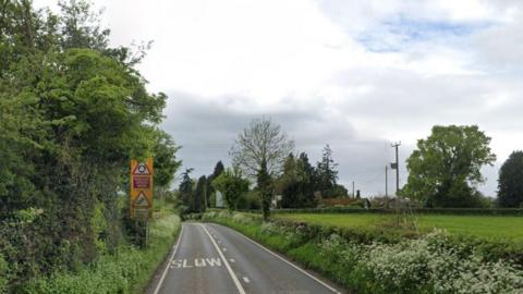 A country road with trees and bushes line each side. Street signs and road markings can be seen in the background.