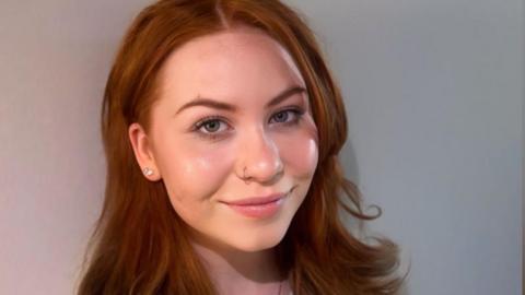 A professional headshot of Liliyana who is smiling to camera. She has long, slightly wavy brown to reddish hair and stud earrings. She is wearing a grey jumper