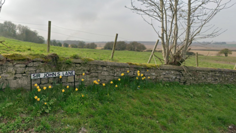 A road sign reading Sir John's Lane in Sherburn in Elmet. The sign is in front of a stone wall about one metre high and is surrounded by daffodils. There are open fields in the background. 