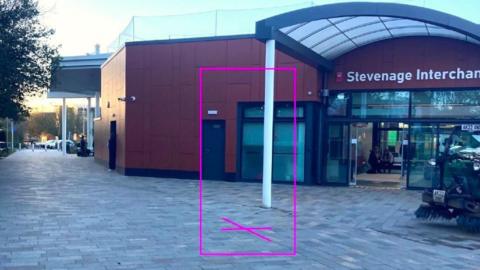 The outside of a bus station, showing a building with a large door and paving outside. There is a pink box, with a cross on the ground, signifying where a statue will be put.