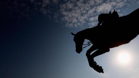 A horse and rider jumping in silhouette against a dark blue sky with high, broken cloud and a hazy white sun.