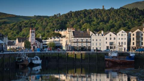 Ramsey quayside overlooking the harbour, buildings like the quay in front of a leafy hill with the Albert Tower above, on a sunny day.