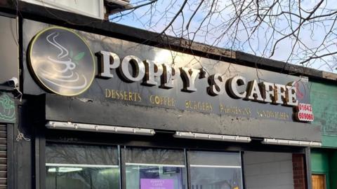 Outside Poppy's Caffe - the name sign above the entrance is in white lettering  - there is also a sign saying Desserts Coffee Burgers Parmesans Sandwiches in yellow. There is a logo to the left of this text, of a white cup of tea with steam rising from the top. The background of the sign is black.