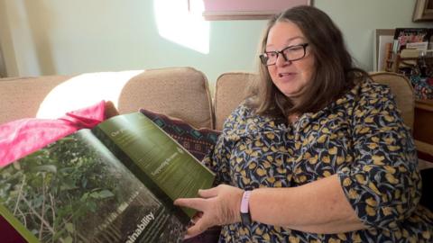 Jane Bertelli, a woman in her mid sixties, sits on the sofa in her home, looking through one of the brochures sent to her by Ethical Forestry Limited.
