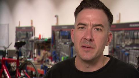 Darrell, a man with dark hair wearing a black t-shirt speaking to the 91ȱ from inside the bike workshop at Pentonville Prison