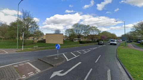 Burn Lane in Newton Aycliffe. The image has been taken from a roundabout and shows a pedestrian island between the two lanes. Brown brick houses stand to the left of the road.