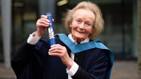 A woman holding a diploma, she is smiling at the camera. She has light hair.