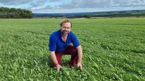 Simon Bainbridge is kneeling down in a field of pasture. He has a short beard and short wavy brown hair.