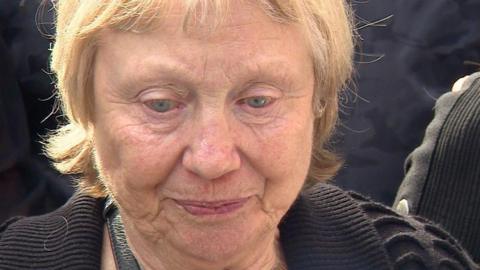 A close-up of an elderly woman with short blonde hair and wearing a black coat.