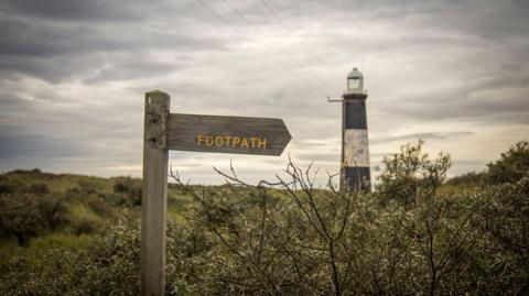 A wooden signpost has yellow text stating 'footpath' on it. A black and white striped lighthouse and the sea can be seen in the background on an overcast day.