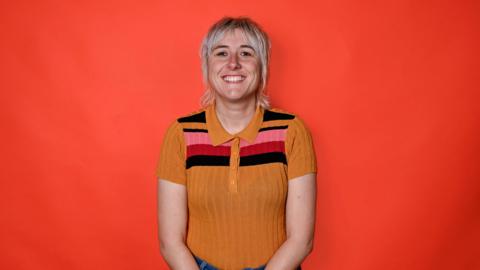 Maisie Adams wearing a yellow striped top with pink and red on it. She has blonde short hair and is standing in front of a red background. 