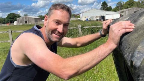 A man wearing a blue vest pushing an old tyre on a grassy field.