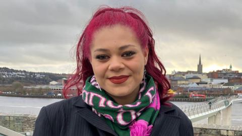 Hannah Richardson, a woman in a black suit with pink hair and a green and pink scarf. Behind her is the River Foyle and the city of Derry with the white Peace Bridge in the background. 