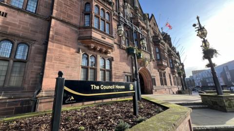 The exterior of the Coventry City Council headquarters is pictured. A sign in the foreground has a black background with gold writing which reads "The Council House". There are stone steps and the building has a sandstone structure with large ornate lead-lined windows. A singular Union Flag flies from one of several flagpoles protruding from the front of the building.