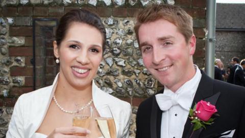 Tom and his late wife Laura smile at the camera whilst holding champagne glasses. Tom wears a black jacket and white bow tie with a pink rose on his lapel,  while Laura wears a white collared dress, pearl necklace and pearl earrings. They stand in front of a stone wall. 