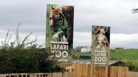 The entrance to South Lakes Safari Zoo. Two giant wooden signs - one picturing a tiger and another a giraffe - stand above a wooden fence. Hills and buildings are in the distance.